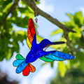 Hummingbird suncatcher in mid-flight with a blue and turquoise body and a fan of rainbow tail feathers, in a hand-shaped soldered metal frame