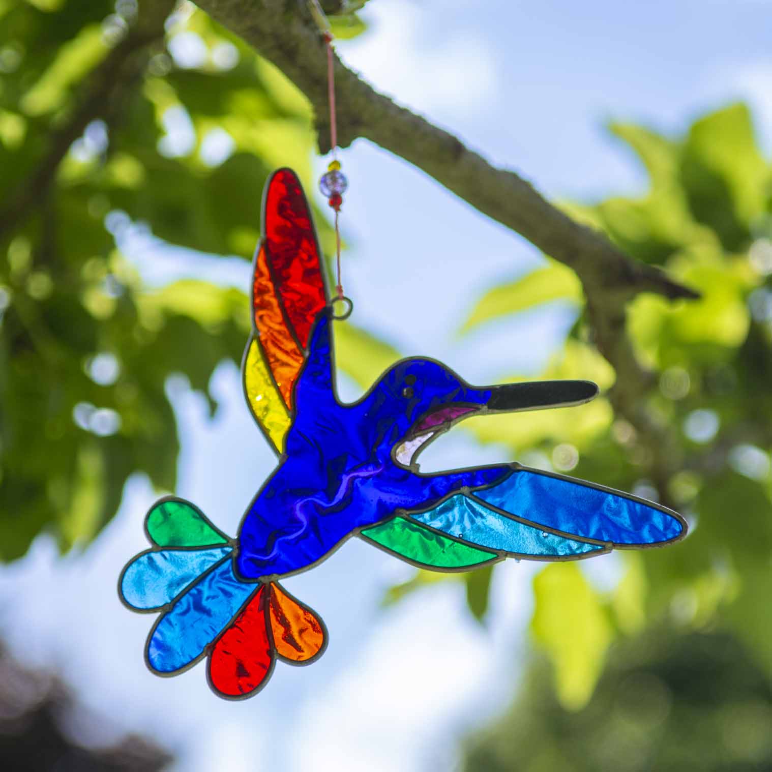 Hummingbird suncatcher in mid-flight with a blue and turquoise body and a fan of rainbow tail feathers, in a hand-shaped soldered metal frame