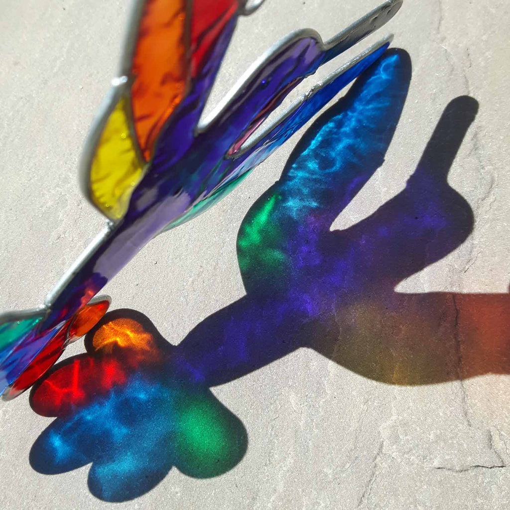 Hummingbird suncatcher in mid-flight with a blue and turquoise body and a fan of rainbow tail feathers, in a hand-shaped soldered metal frame