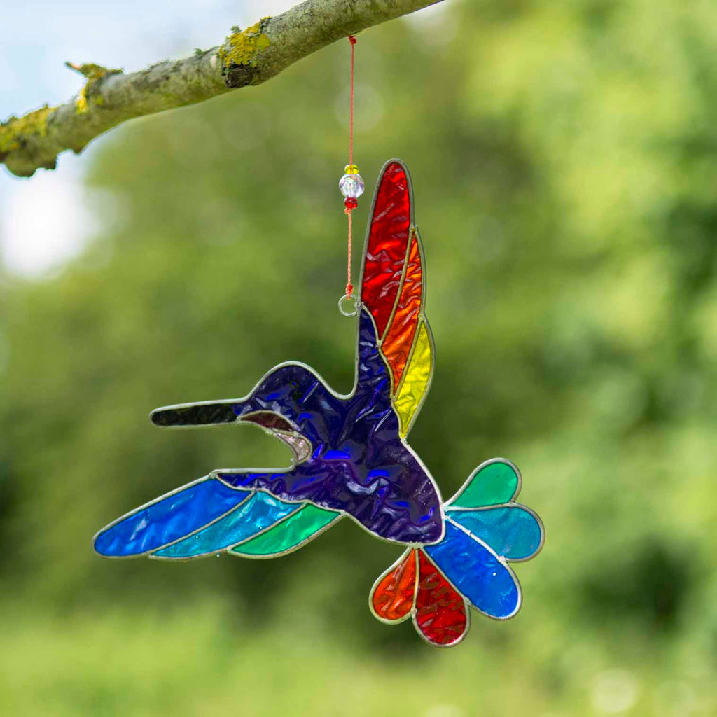 Hummingbird suncatcher in mid-flight with a blue and turquoise body and a fan of rainbow tail feathers, in a hand-shaped soldered metal frame