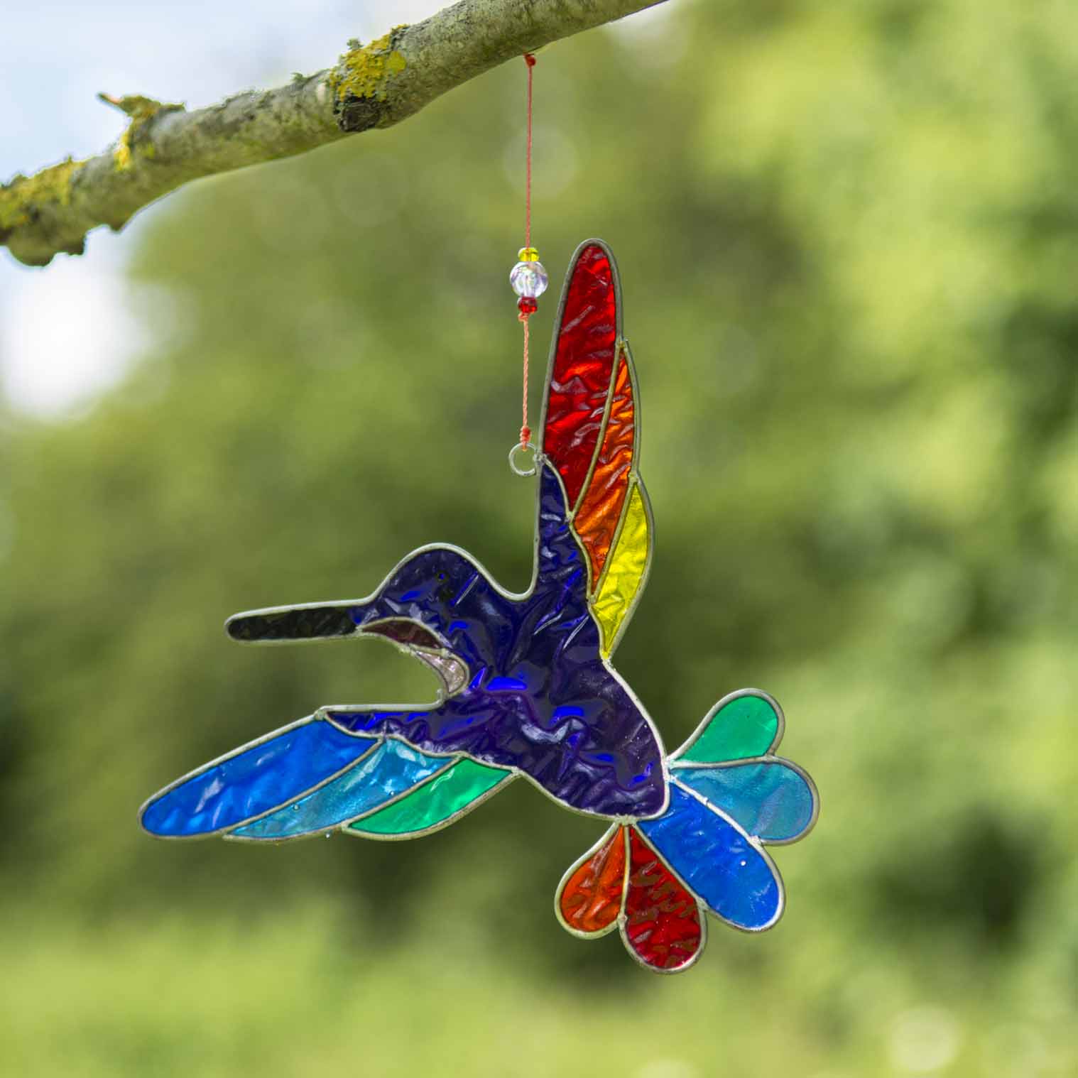 Hummingbird suncatcher in mid-flight with a blue and turquoise body and a fan of rainbow tail feathers, in a hand-shaped soldered metal frame