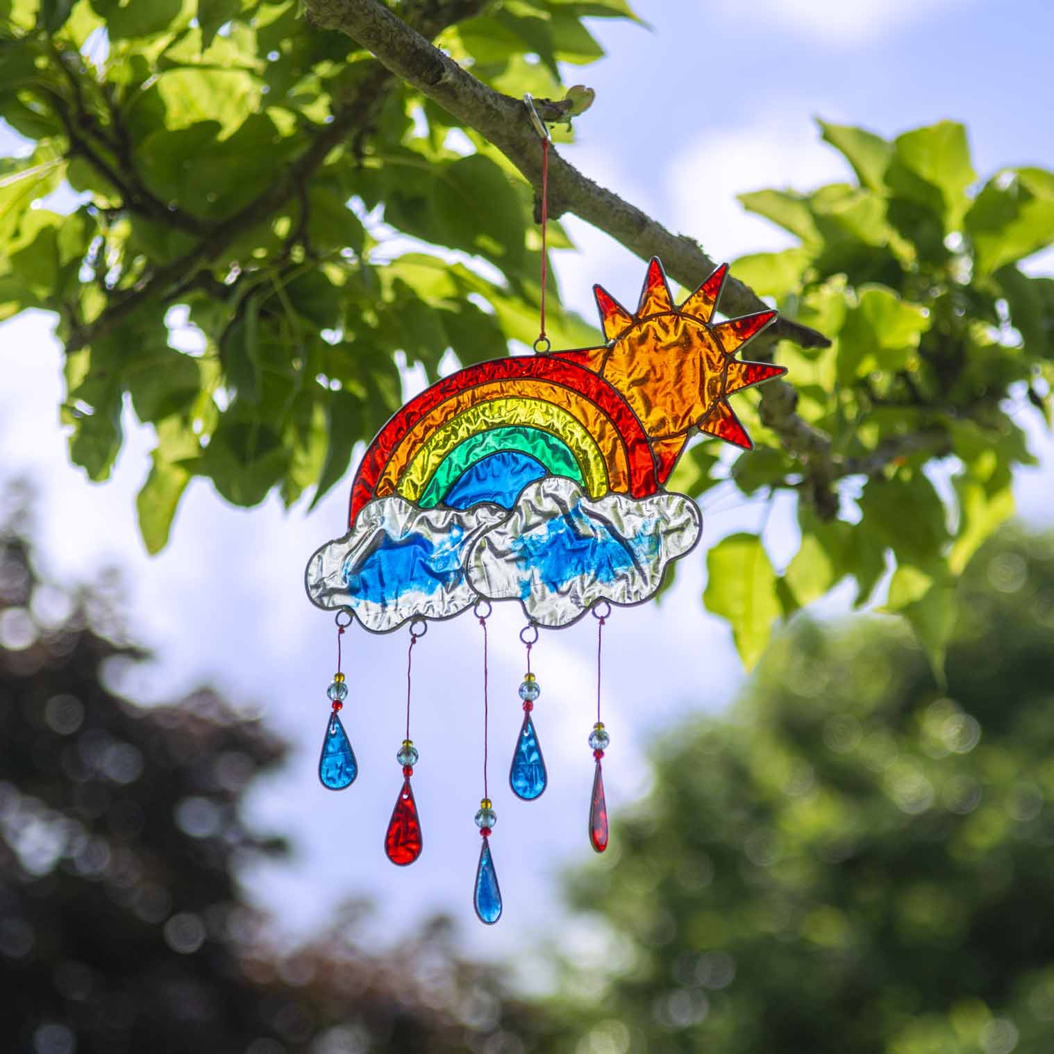 Stained-glass effect suncatcher of a red sun rising over a rainbow arc, with two clouds and dangling red, yellow and blue raindrop pendants