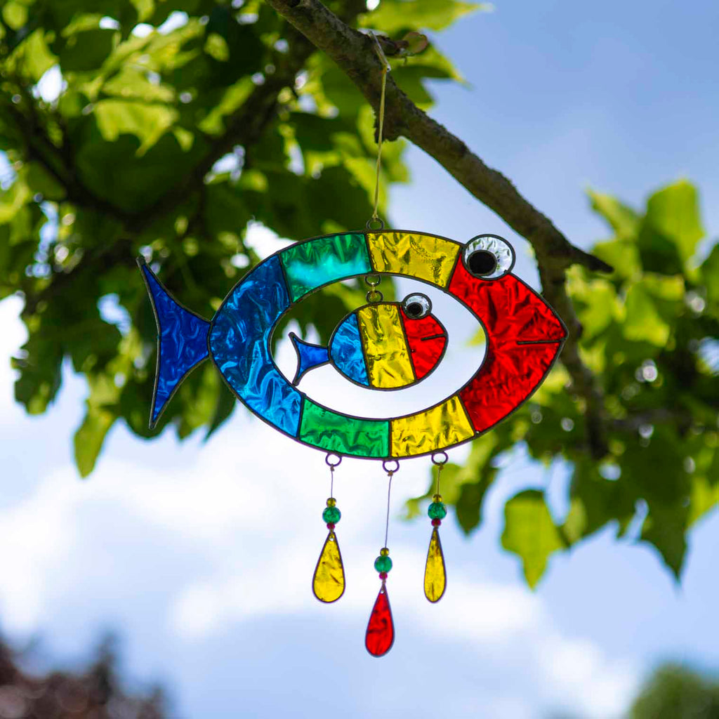 Suncatcher of two cheerful fish, a big one with a smaller fish inside, in bright primary colours with red, yellow and amber glass teardrops dangling beneath