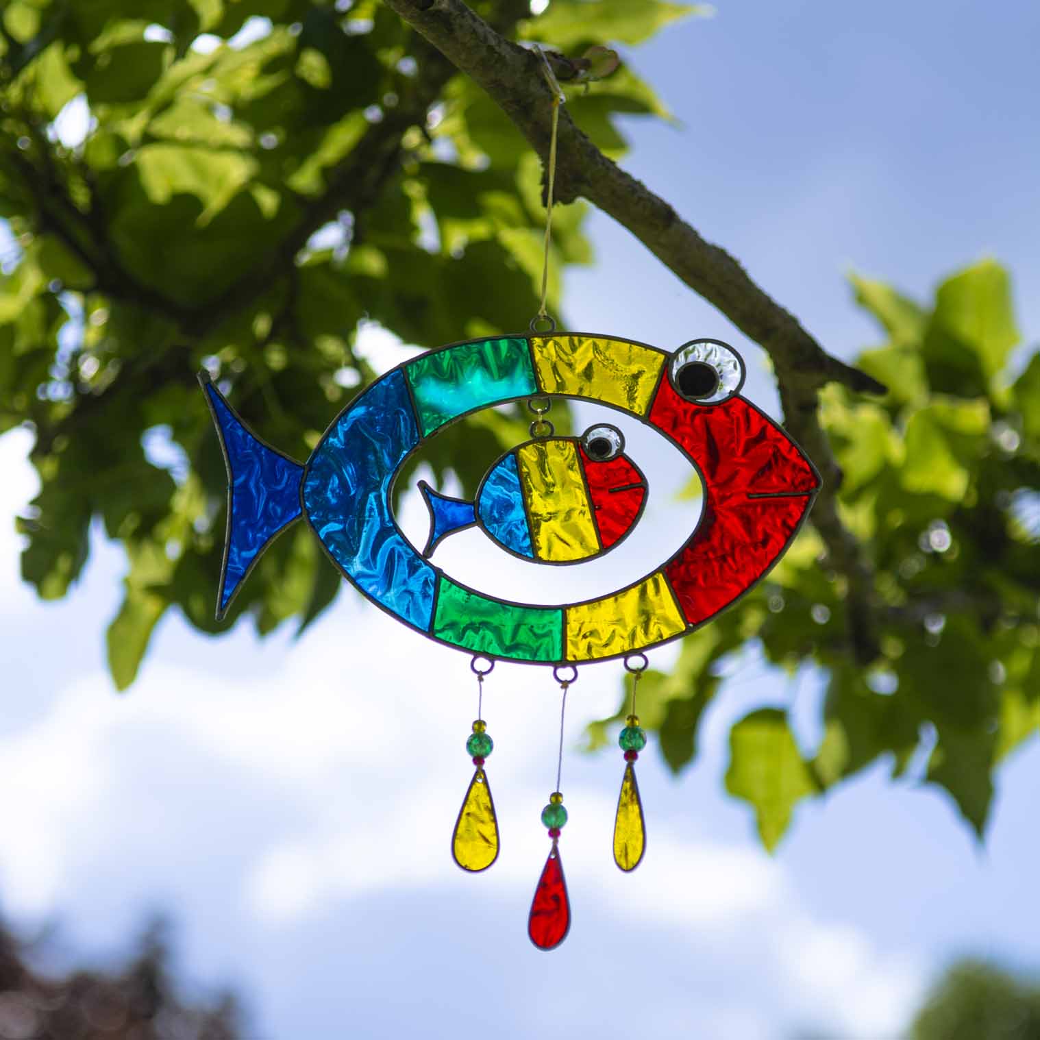 Suncatcher of two cheerful fish, a big one with a smaller fish inside, in bright primary colours with red, yellow and amber glass teardrops dangling beneath