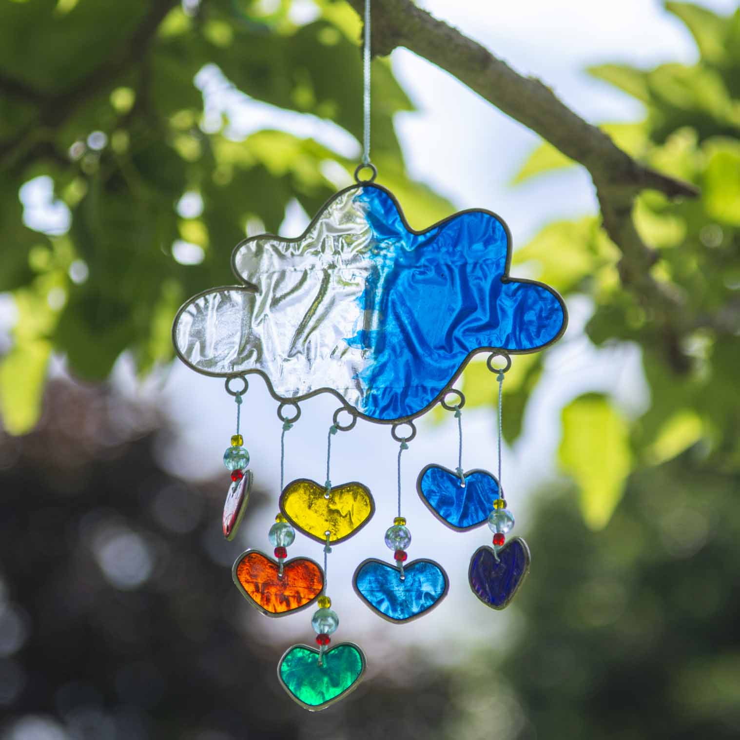 Blue resin cloud suncatcher with six threads of rainbow hearts dangling beneath in jewel tones of red, orange, yellow, green, blue and purple