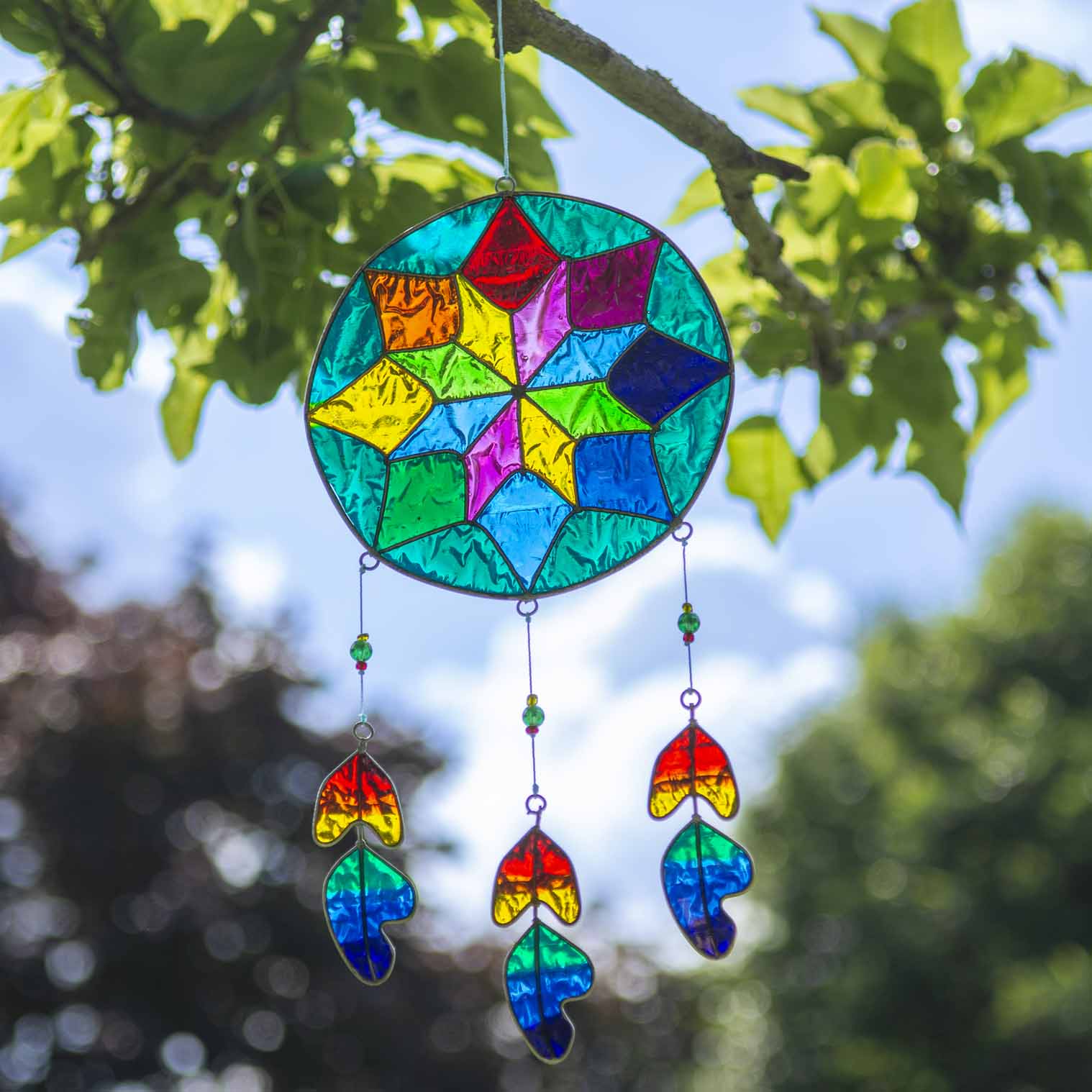 Round dreamcatcher-style suncatcher with a geometric resin web in blue, green, ruby and amber, with three rainbow feather pieces dangling beneath