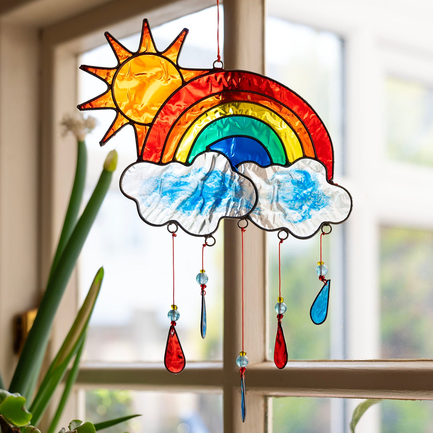 Stained-glass effect suncatcher of a red sun rising over a rainbow arc, with two clouds and dangling red, yellow and blue raindrop pendants